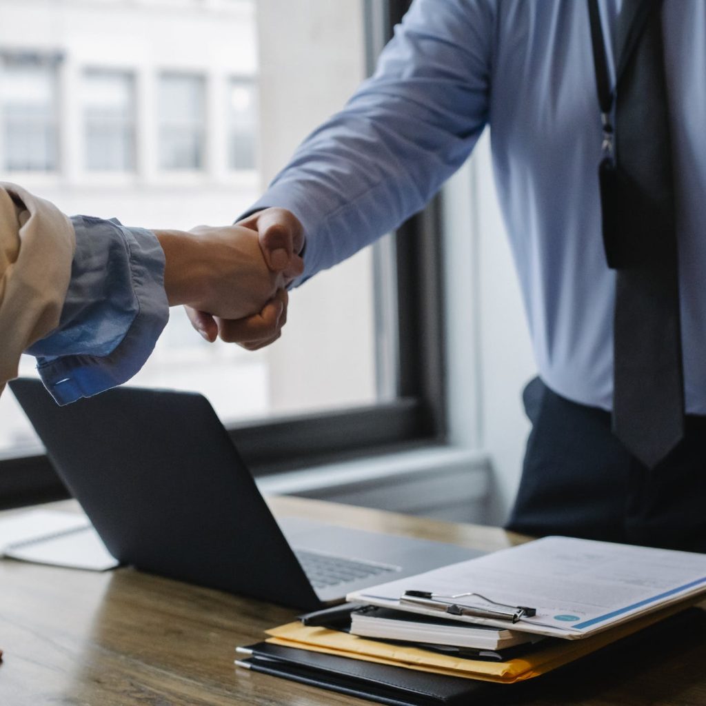 Crop unrecognizable coworkers in formal wear standing at table with laptop and documents while greeting each other before meeting